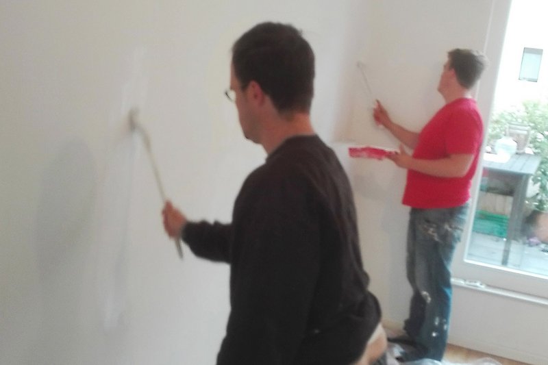 Zwei junge Männer stehen vor einer leeren Wand und streichen sie mit Pinsel und Rolle mit weißer Farbe.