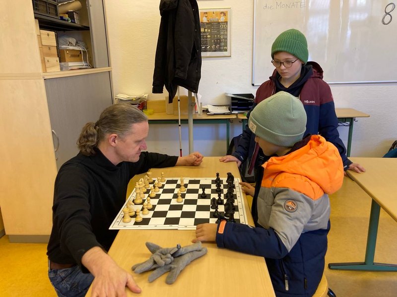 Zwei Kinder und ein Mann beim Schachspielen. Im HIntergrund lehnt ein Blinderstock an der Wand. Zwei Kinder und ein Mann beim Schachspielen. Im Hintergrund lehnt ein Blindenstock an der Wand. Der Mann und ein Kind hocken vor einem Tisch mit einem Schachbrett und Figuren darauf. Das 2. Kind steht hinter dem Tisch.