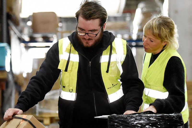 Bei der Arbeit im Lager mit Verpackungen Ein junger Mann und eine junge Frau mit gelben Warnwesten stehen in einem Lager mit Kartons und Verpackungen. Beide schauen auf einen Karton, der angeschnitten links im unteren Bildrand zu sehen ist.